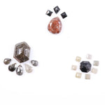 Assorted natural fancy color diamonds including salt and pepper hexagon cut, pear rose cuts, peach oval, dark geometric step cut center stone, and small pyramid-cut accent diamonds arranged on white background.