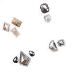 Assorted natural salt and pepper diamonds in geometric fancy cuts including kite, shield, emerald, pear, half moon, and octagon shapes in earthy tones of grey, champagne, cognac, and blush with visible natural inclusions.