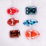 Six fancy color lab grown diamonds displayed on a white background, including pear, marquise, emerald and oval cuts in vibrant pink, blue and cognac tones, precision faceted for fine jewelry settings.