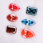 Six fancy color lab grown diamonds displayed on a white background, including pear, marquise, emerald and oval cuts in vibrant pink, blue and cognac tones, precision faceted for fine jewelry settings.
