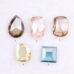 Five lab grown diamonds in various shapes including pear, cushion, and square step cut, displayed in pastel shades of pink, champagne, yellow, and blue on a white background.
