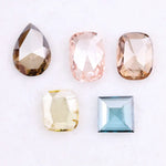 Five lab grown diamonds in various shapes including pear, cushion, and square step cut, displayed in pastel shades of pink, champagne, yellow, and blue on a white background.
