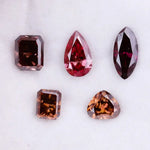 Five lab grown fancy color diamonds in red and cognac tones featuring pear, emerald, marquise, and trillion cuts displayed on a white background, loose sustainable gemstones for custom fine jewelry.