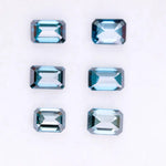 Six emerald cut lab grown blue diamonds in teal and deep ocean blue tones arranged on a white background, showcasing step cut facets and rectangular shapes for fine jewelry and engagement ring settings.