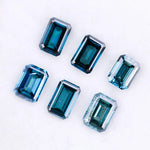 Six emerald cut lab grown blue diamonds in teal and deep ocean blue tones arranged on a white background, showcasing step cut facets and rectangular shapes for fine jewelry and engagement ring settings.