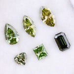 Six fancy color lab grown diamonds in green and yellow shades displayed on white background, featuring pear, marquise, heart, princess, and emerald cuts.