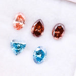 Five loose lab grown diamonds displayed on a white background, featuring three pink diamonds in oval, pear, and round cuts, alongside two vivid blue diamonds in heart and oval shapes, showcasing brilliant faceting and high clarity.