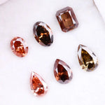 Six fancy colored lab grown diamonds in pink, peach, and cognac tones featuring emerald cut, oval cut, and pear cut shapes displayed on a white background, ethical CVD loose stones for custom fine jewelry.