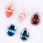 Collection of fancy color lab grown diamonds including pink marquise cut, light peach marquise, brown pear shape, blue oval cut and blue pear cut loose diamonds displayed on white background
