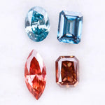 Four fancy color lab grown diamonds including oval and emerald cut blue stones and marquise and radiant cut red gemstones displayed on a white background.