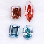 Four fancy color lab grown diamonds including oval and emerald cut blue stones and marquise and radiant cut red gemstones displayed on a white background.