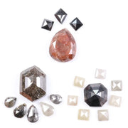 Assorted natural fancy color diamonds including salt and pepper hexagon cut, pear rose cuts, peach oval, dark geometric step cut center stone, and small pyramid-cut accent diamonds arranged on white background.