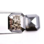 Pair of emerald cut salt and pepper lab grown diamonds with black and white inclusions in geometric step cut, shown loose on white background.