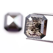 Pair of emerald cut salt and pepper lab grown diamonds with black and white inclusions in geometric step cut, shown loose on white background.