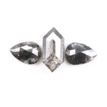 Three natural salt and pepper diamonds including a kite cut center stone and two pear shaped side stones with grey inclusions and rose cut faceting on a white background.