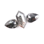 Three natural salt and pepper diamonds including a kite cut center stone and two pear shaped side stones with grey inclusions and rose cut faceting on a white background.
