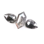 Three natural salt and pepper diamonds including a kite cut center stone and two pear shaped side stones with grey inclusions and rose cut faceting on a white background.