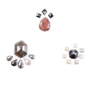 Assorted natural fancy color diamonds including salt and pepper hexagon cut, pear rose cuts, peach oval, dark geometric step cut center stone, and small pyramid-cut accent diamonds arranged on white background.