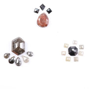 Assorted natural fancy color diamonds including salt and pepper hexagon cut, pear rose cuts, peach oval, dark geometric step cut center stone, and small pyramid-cut accent diamonds arranged on white background.