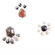Assorted natural fancy color diamonds including salt and pepper hexagon cut, pear rose cuts, peach oval, dark geometric step cut center stone, and small pyramid-cut accent diamonds arranged on white background.
