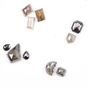 Assorted natural salt and pepper diamonds in geometric fancy cuts including kite, shield, emerald, pear, half moon, and octagon shapes in earthy tones of grey, champagne, cognac, and blush with visible natural inclusions.