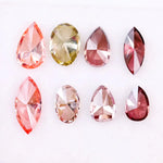 Eight faceted lab grown colored diamonds in pear, oval, and marquise cuts featuring fancy pink, yellow, and deep red tones arranged on a white background.