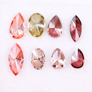 Eight faceted lab grown colored diamonds in pear, oval, and marquise cuts featuring fancy pink, yellow, and deep red tones arranged on a white background.