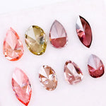 Eight faceted lab grown colored diamonds in pear, oval, and marquise cuts featuring fancy pink, yellow, and deep red tones arranged on a white background.