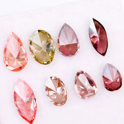 Eight faceted lab grown colored diamonds in pear, oval, and marquise cuts featuring fancy pink, yellow, and deep red tones arranged on a white background.