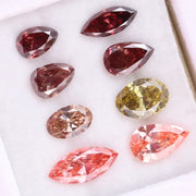 Eight faceted lab grown colored diamonds in pear, oval, and marquise cuts featuring fancy pink, yellow, and deep red tones arranged on a white background.