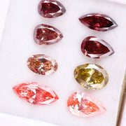 Eight faceted lab grown colored diamonds in pear, oval, and marquise cuts featuring fancy pink, yellow, and deep red tones arranged on a white background.