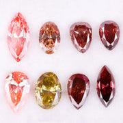 Eight faceted lab grown colored diamonds in pear, oval, and marquise cuts featuring fancy pink, yellow, and deep red tones arranged on a white background.