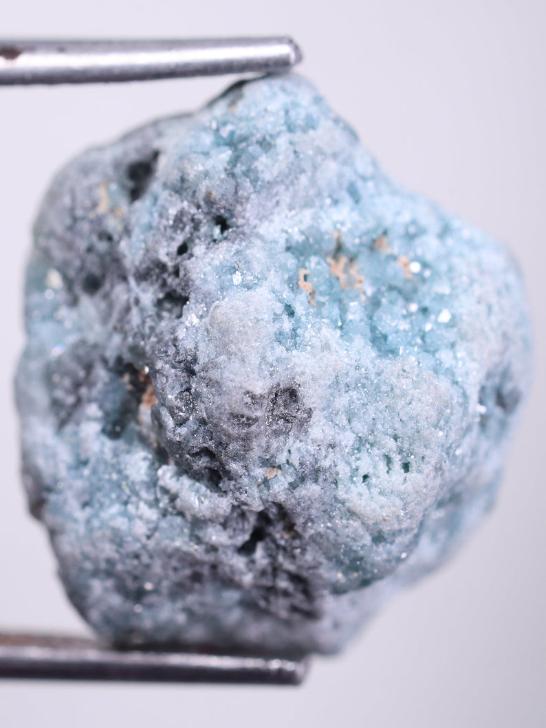 Blue natural diamond rock held by tweezers on a light gray background