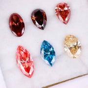 Six fancy color lab grown diamonds on white background including yellow oval cut, blue marquise cut, pink marquise cut, peach pear cut, deep red pear cut, and red oval cut brilliant stones for custom jewelry design.