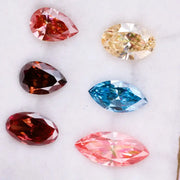 Six fancy color lab grown diamonds on white background including yellow oval cut, blue marquise cut, pink marquise cut, peach pear cut, deep red pear cut, and red oval cut brilliant stones for custom jewelry design.