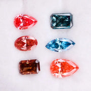 Six fancy color lab grown diamonds displayed on a white background, including pear, marquise, emerald and oval cuts in vibrant pink, blue and cognac tones, precision faceted for fine jewelry settings.