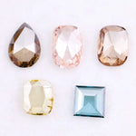 Five lab grown diamonds in various shapes including pear, cushion, and square step cut, displayed in pastel shades of pink, champagne, yellow, and blue on a white background.