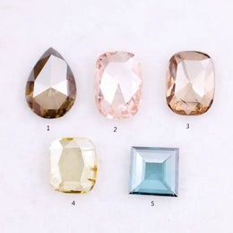 Five lab grown diamonds in various shapes including pear, cushion, and square step cut, displayed in pastel shades of pink, champagne, yellow, and blue on a white background.