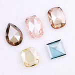 Five lab grown diamonds in various shapes including pear, cushion, and square step cut, displayed in pastel shades of pink, champagne, yellow, and blue on a white background.