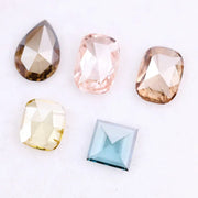 Five lab grown diamonds in various shapes including pear, cushion, and square step cut, displayed in pastel shades of pink, champagne, yellow, and blue on a white background.