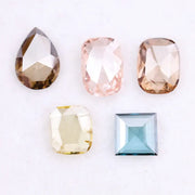 Five lab grown diamonds in various shapes including pear, cushion, and square step cut, displayed in pastel shades of pink, champagne, yellow, and blue on a white background.