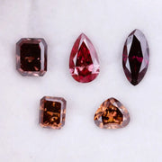 Five lab grown fancy color diamonds in red and cognac tones featuring pear, emerald, marquise, and trillion cuts displayed on a white background, loose sustainable gemstones for custom fine jewelry.