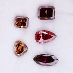 Five lab grown fancy color diamonds in red and cognac tones featuring pear, emerald, marquise, and trillion cuts displayed on a white background, loose sustainable gemstones for custom fine jewelry.