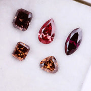 Five lab grown fancy color diamonds in red and cognac tones featuring pear, emerald, marquise, and trillion cuts displayed on a white background, loose sustainable gemstones for custom fine jewelry.