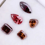 Five lab grown fancy color diamonds in red and cognac tones featuring pear, emerald, marquise, and trillion cuts displayed on a white background, loose sustainable gemstones for custom fine jewelry.