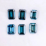 Six emerald cut lab grown blue diamonds in teal and deep ocean blue tones arranged on a white background, showcasing step cut facets and rectangular shapes for fine jewelry and engagement ring settings.