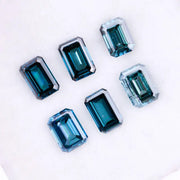 Six emerald cut lab grown blue diamonds in teal and deep ocean blue tones arranged on a white background, showcasing step cut facets and rectangular shapes for fine jewelry and engagement ring settings.