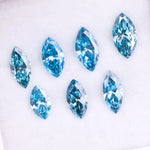 Seven marquise cut blue lab grown diamonds in vivid teal and aqua shades displayed on a white background, showcasing brilliant faceting and elongated shapes for engagement rings or custom jewelry.