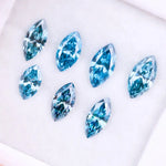 Seven marquise cut blue lab grown diamonds in vivid teal and aqua shades displayed on a white background, showcasing brilliant faceting and elongated shapes for engagement rings or custom jewelry.