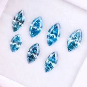 Seven marquise cut blue lab grown diamonds in vivid teal and aqua shades displayed on a white background, showcasing brilliant faceting and elongated shapes for engagement rings or custom jewelry.